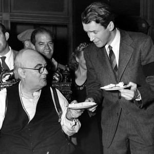 IT'S A WONDERFUL LIFE, James Stewart eating cake with Lionel Barrymore on the set, director Frank Capra in background, 1946